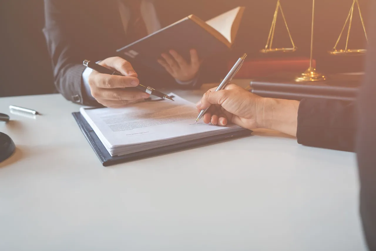 Person signing documents at a desk beside a set of brass scales